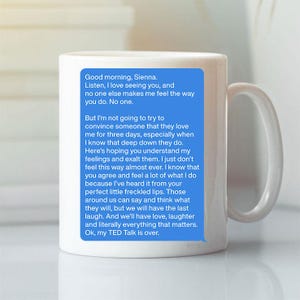 Könnte beinhalten: Weiße Keramik-Tasse mit gebogenem Henkel. Auf der Vorderseite befindet sich eine blaue Sprechblase mit einer Nachricht. Die Nachricht beginnt mit "Good morning, Sienna" und setzt sich mit einer persönlichen Nachricht fort.