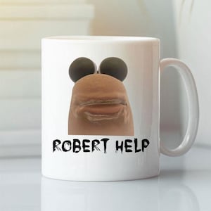Könnte beinhalten: Weiße Keramik-Tasse mit einem humorvollen Design. Sie zeigt ein Cartoon-Gesicht mit schwarzen Augen und einer braunen Nase und Mund. Der Text "ROBERT HELP" ist in schwarzen, stilisierten Buchstaben darunter gedruckt.