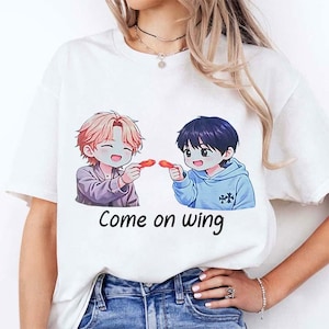 Könnte beinhalten: Weißes T-Shirt mit einer Cartoon-Illustration von zwei Anime-Charakteren, die rote Objekte halten. Die Charaktere lächeln, und der Text "Come on wing" ist darunter in Schwarz gedruckt.