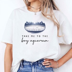 Pode incluir: T-shirt branca com um gráfico azul e branco de um estádio e o texto "TAKE ME TO THE boy aquarium" em escrita preta. A camisa é feita de um material macio.