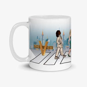 May include: White ceramic mug featuring an illustration of a cityscape with the text "THE VIEW" in gold. The mug depicts three figures walking on a crosswalk. The background includes skyscrapers and a blue sky.