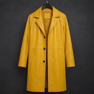 Puede incluir: Un abrigo de cuero amarillo mostaza con un diseño clásico, con solapas, mangas largas y cierre de botones. El abrigo tiene dos bolsillos delanteros y está colgado de una percha negra sobre un fondo oscuro.