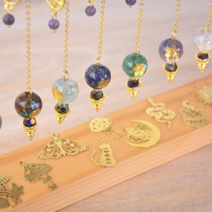 Può includere: Una collezione di pendoli dorati con varie sfere di pietre preziose e cristalli appese a catene. I pendoli sono esposti su una superficie di legno con ciondoli di metallo decorativi.