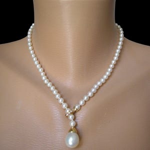 May include: A necklace featuring a strand of small, round, white pearls. The strand connects to a gold-toned metal accent piece encrusted with small clear stones. A single, larger, teardrop-shaped white pearl hangs from a gold-toned metal cap.