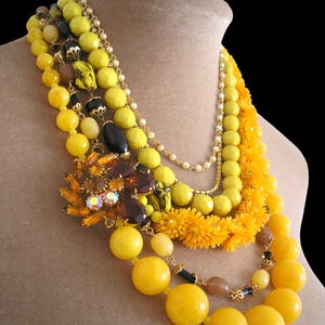 May include: A multi-strand statement necklace featuring various shades of yellow and black beads, including round, faceted, and flower-shaped components. A large, dark amber-coloured floral brooch with iridescent accents serves as a central focal point.