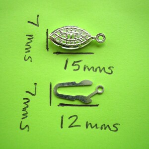 May include: Two silver metal jewelry findings. The first is an oval-shaped clasp with a decorative design, measuring 15 mm long and 7 mm wide. The second is a hook clasp measuring 12 mm long and 7 mm wide.
