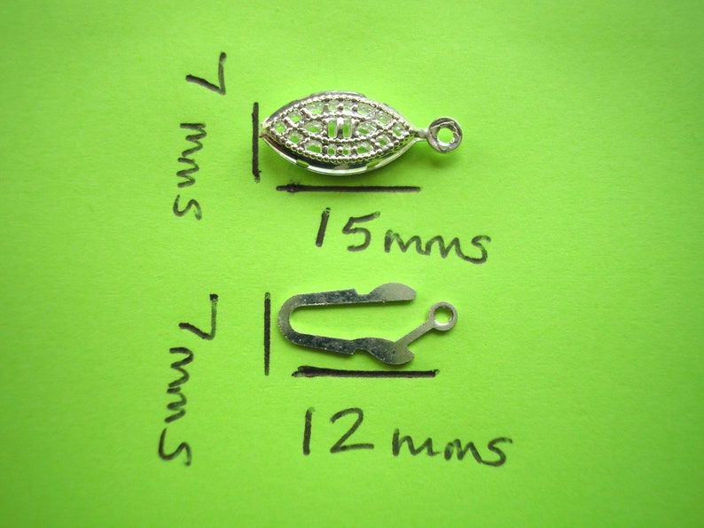May include: Two silver metal jewelry findings. The first is an oval-shaped clasp with a decorative design, measuring 15 mm long and 7 mm wide. The second is a hook clasp measuring 12 mm long and 7 mm wide.
