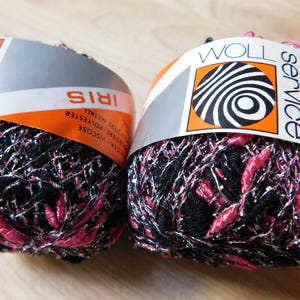 Puede incluir: Dos ovillos de hilo Iris, con hilos negros, rosas y plateados. El hilo está envuelto con etiquetas que dicen "Iris" y "Wollservice". Las etiquetas tienen detalles en naranja y plata.