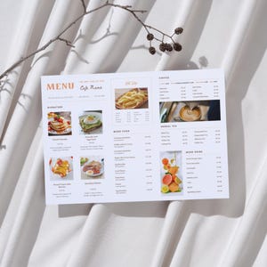 Puede incluir: Un menú de cafetería rectangular impreso en papel blanco con el título MENU, Cafe Name, y secciones para Signature, More Food, Coffee, Herbal Tea y More Drink. El diseño incluye fotografías de tortitas, tostadas de aguacate y frutas.