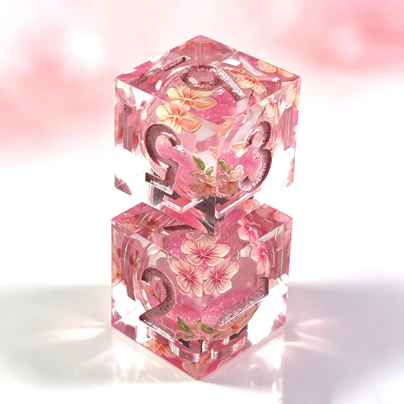 May include: Two stacked translucent resin dice featuring a clear finish with embedded pink cherry blossom floral patterns and fine pink glitter. The dice have large, deeply engraved numbers in a metallic pink finish, showing digits 2, 3, and 9.