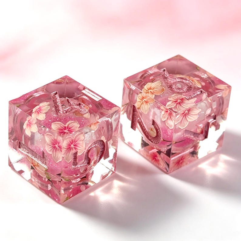 May include: Two transparent resin dice featuring embedded pink cherry blossom flowers and glitter. Each cube has recessed numbers filled with a shimmering pink material, showcasing a clear, glossy finish with floral details visible throughout the interior.