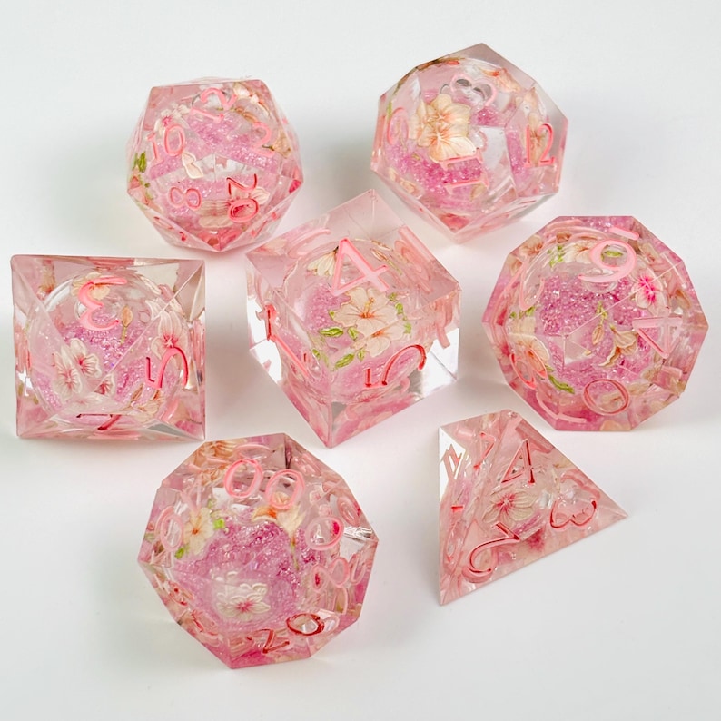 May include: Set of seven polyhedral dice with a floral design. The dice are clear with pink accents and embedded flowers. Each die has pink numbers. The dice are for tabletop games.