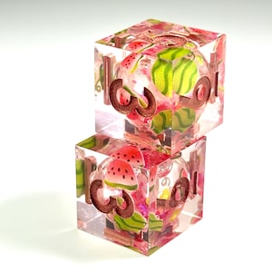 May include: Two stacked transparent resin dice featuring embedded miniature watermelon slices and green rind patterns. The dice have copper-colored numbers engraved on the faces, with the number three clearly visible on the front of both cubes.