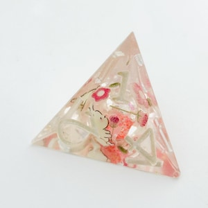 Peut inclure: D&eacute; de jeu triangulaire en r&eacute;sine transparente, orn&eacute; de fleurs roses et d'un petit personnage de dessin anim&eacute; blanc. Le d&eacute; comporte des chiffres blancs et est pos&eacute; sur un fond blanc. Un objet unique pour les jeux de soci&eacute;t&eacute;.