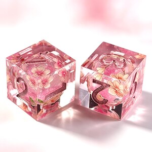 May include: Two six-sided dice made of clear resin, each containing embedded pink cherry blossom illustrations and pink glitter. The dice feature large, recessed numbers in a metallic rose gold finish, with the numbers 2, 3, 5, and 6 visible.