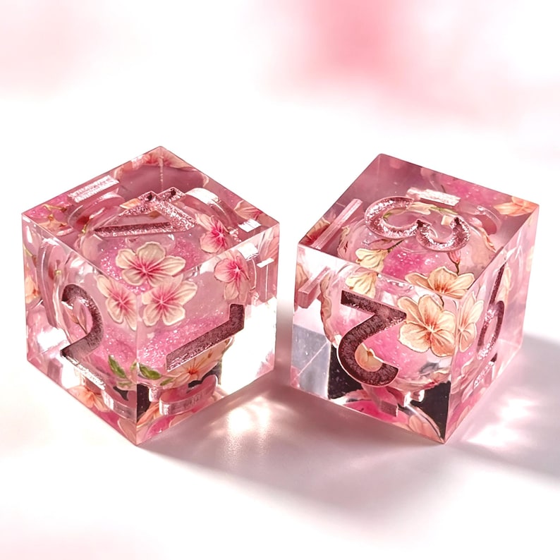 May include: Two six-sided dice made of clear resin, each containing embedded pink cherry blossom illustrations and pink glitter. The dice feature large, recessed numbers in a metallic rose gold finish, with the numbers 2, 3, 5, and 6 visible.
