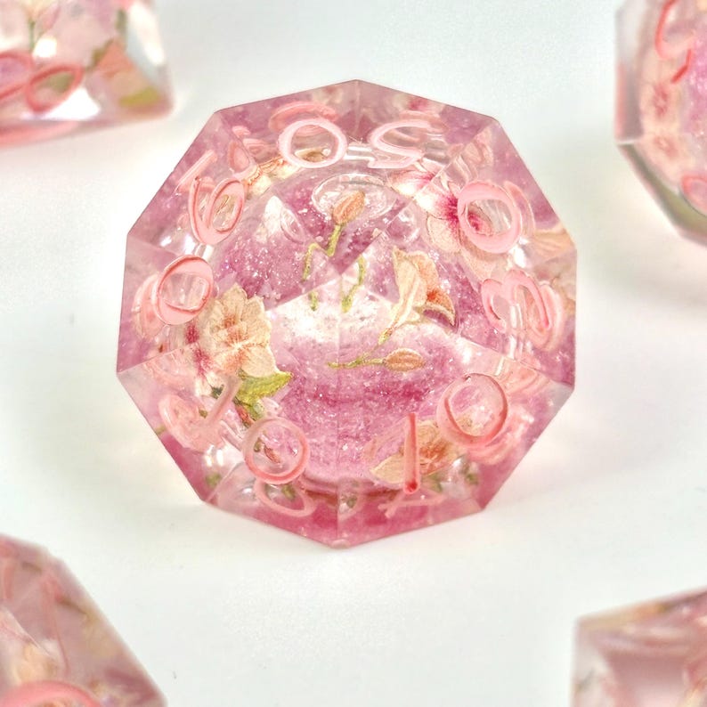 May include: A set of polyhedral dice with a floral design. The dice are a translucent pink with embedded flowers and pink numbering. The dice are designed for tabletop games.