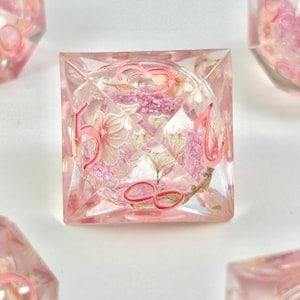May include: A clear, square-shaped die with pink numbers and floral inclusions. The die features a delicate arrangement of pink and white flowers encased within the transparent resin. The numbers are painted in a matching pink hue.