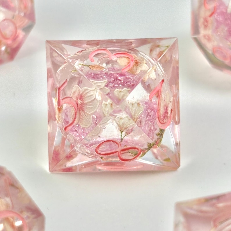 May include: A clear, square-shaped die with pink numbers and floral inclusions. The die features a delicate arrangement of pink and white flowers encased within the transparent resin. The numbers are painted in a matching pink hue.