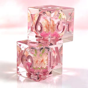 May include: Two clear resin dice stacked vertically, each containing a spherical core with pink floral patterns and green leaves. The dice feature metallic pink numbers embedded within the transparent cube structure, showcasing a detailed botanical design.