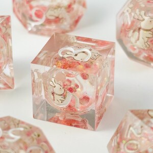 Peut inclure: D&eacute;s en r&eacute;sine transparente de couleur rose. Chaque d&eacute; pr&eacute;sente une illustration de lapin blanc avec un n&oelig;ud rose et un bouquet de roses roses. Les d&eacute;s sont de forme cubique avec un int&eacute;rieur sph&eacute;rique.