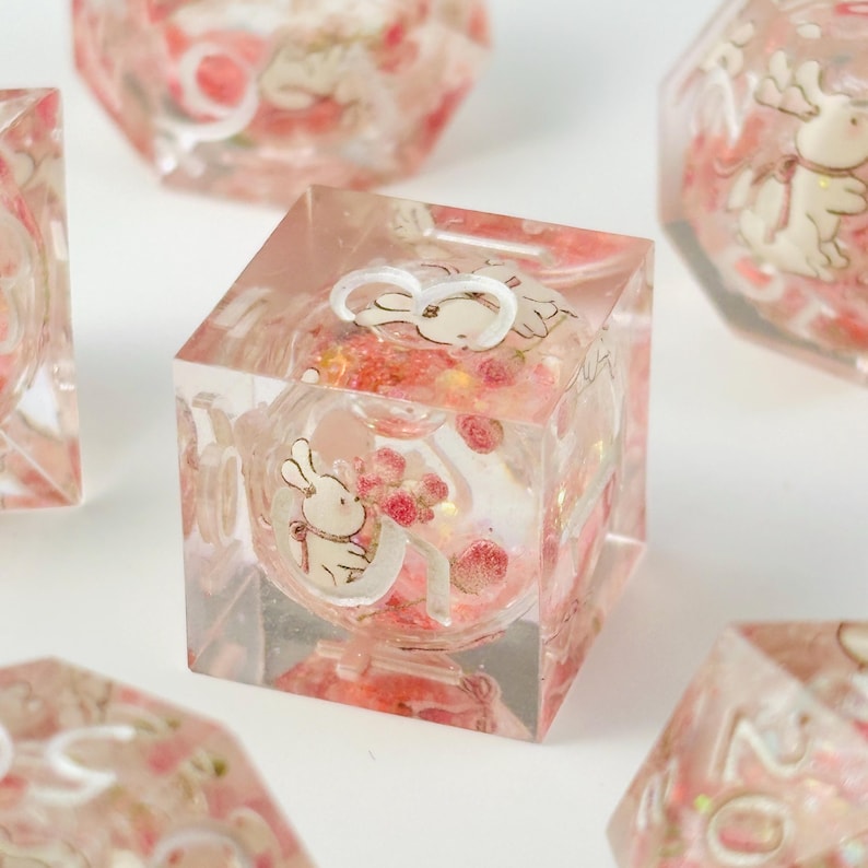 Peut inclure: D&eacute;s en r&eacute;sine transparente de couleur rose. Chaque d&eacute; pr&eacute;sente une illustration de lapin blanc avec un n&oelig;ud rose et un bouquet de roses roses. Les d&eacute;s sont de forme cubique avec un int&eacute;rieur sph&eacute;rique.