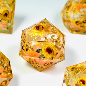 May include: A multi-faceted polyhedral die made of clear resin containing embedded miniature yellow sunflower blossoms and iridescent glitter particles. The die features engraved numbers filled with a metallic gold pigment.