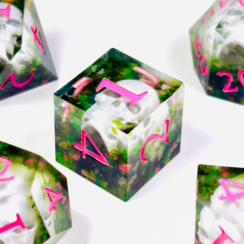 Op de afbeelding: Set polyhedrale dobbelstenen met een schedel in elke dobbelsteen. De dobbelstenen zijn transparant met een bloemenpatroon en felroze cijfers. De schedel is wit. Dobbelstenen voor tabletop games.