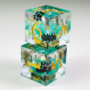 May include: Two stacked translucent resin dice featuring a clear exterior and a teal-colored interior filled with small glittery particles. Each die contains black cat silhouettes and gold-colored numbers, including a prominent six and one.