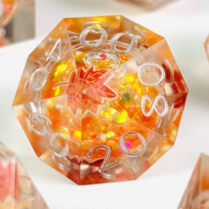 May include: A large, multi-sided die with an orange and yellow interior, featuring a maple leaf design. The die has silver-colored numbers and is made of a clear, resin-like material. The overall theme is autumn or fall.