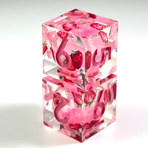 May include: Two stacked clear resin dice featuring embedded strawberry imagery and pink glitter accents. The dice have deep pink engraved numbers, including a prominent six and five on the top die, and a seven and six on the bottom die, with clear edges.