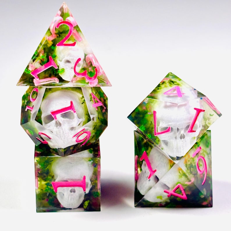 Op de afbeelding: Set unieke, handgemaakte dobbelstenen voor tabletop games. Elke dobbelsteen heeft een witte schedel in heldere hars met roze cijfers en bloemaccenten. De dobbelstenen hebben verschillende geometrische vormen, waaronder een piramide en een kubus.