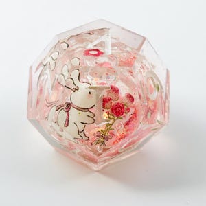 Peut inclure: D&eacute; de r&eacute;sine clair &agrave; multiples facettes avec un design fantaisiste. &Agrave; l'int&eacute;rieur, un lapin blanc avec un n&oelig;ud rose et un bouquet de roses rouges sont visibles. Le d&eacute; est rempli de paillettes roses et porte le chiffre 1.
