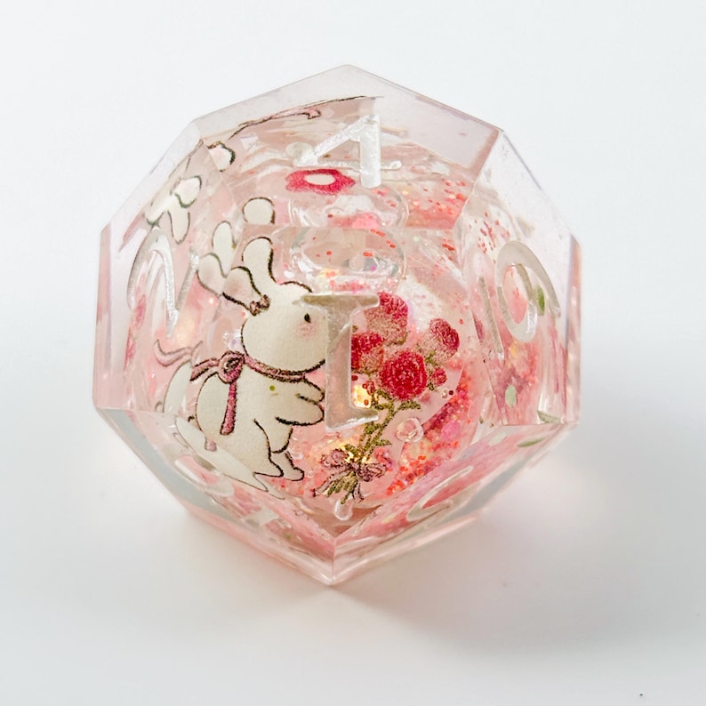 Peut inclure: D&eacute; de r&eacute;sine clair &agrave; multiples facettes avec un design fantaisiste. &Agrave; l'int&eacute;rieur, un lapin blanc avec un n&oelig;ud rose et un bouquet de roses rouges sont visibles. Le d&eacute; est rempli de paillettes roses et porte le chiffre 1.