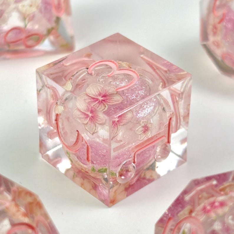 May include: A clear, cube-shaped resin die with a pink floral design and the number 3. The die is filled with glitter and small pink flowers. The edges of the die are outlined in pink.