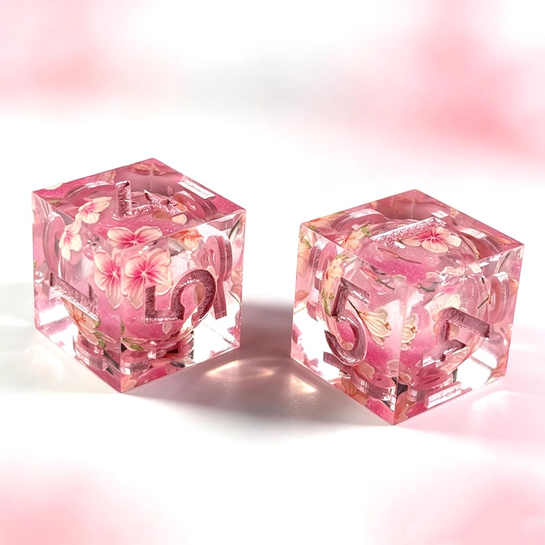 May include: Two six-sided dice made of clear resin with a pink tint. Each die contains embedded pink and white flower petals and fine glitter. The numbers are engraved into the surface of the dice and filled with a metallic pink pigment.