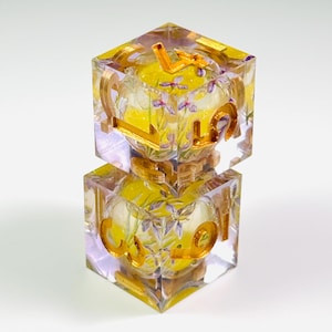 May include: Two stacked translucent resin dice featuring embedded purple floral patterns and bright yellow centers. Each die has gold-colored metallic numbers embossed on the exterior faces, including visible digits like four, five, three, and nine.