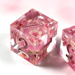May include: A translucent pink resin cube die featuring embedded cherry blossom illustrations and glittery pink accents. The die has large, recessed numbers with a metallic finish, including a visible number two on the front face.