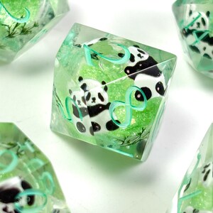 May include: Clear, green-tinted dice with panda bear designs. The dice feature black and white panda illustrations and light green numbers. The dice are set against a green, textured background, with a few other dice visible in the background.