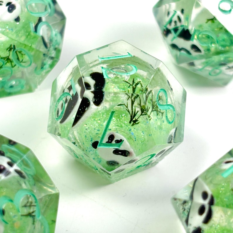 May include: Several polyhedral dice with a panda bear theme. The dice are clear with a green glitter background and panda bear images. The numbers are in a teal color. Bamboo stalks are also visible inside the dice.