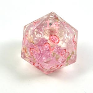 May include: A clear, multi-sided die with pink floral inclusions and pink numbering. The die is filled with small pink glitter and dried flowers. The number 14 is visible on the top face. A unique and decorative gaming accessory.