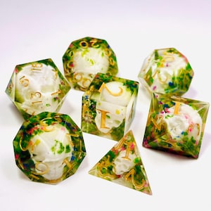 Op de afbeelding: Set van zeven polyhedrale dobbelstenen met een schedelontwerp in heldere hars. De dobbelstenen hebben gouden cijfers en zijn versierd met kleurrijke bloemen. Perfect voor tabletop games.