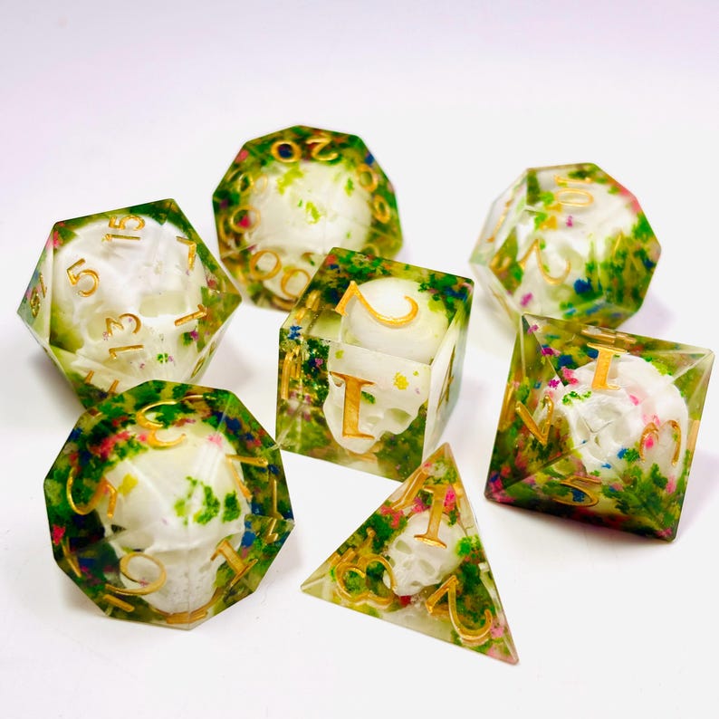 Op de afbeelding: Set van zeven polyhedrale dobbelstenen met een schedelontwerp in heldere hars. De dobbelstenen hebben gouden cijfers en zijn versierd met kleurrijke bloemen. Perfect voor tabletop games.