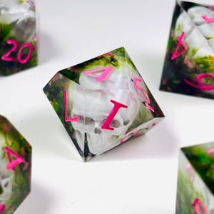 Op de afbeelding: Set polyhedrale dobbelstenen met een schedelontwerp. De dobbelstenen zijn helder met een schedel en bloemeninsluitsels, en felroze cijfers. De dobbelstenen zijn voor tabletopspellen.