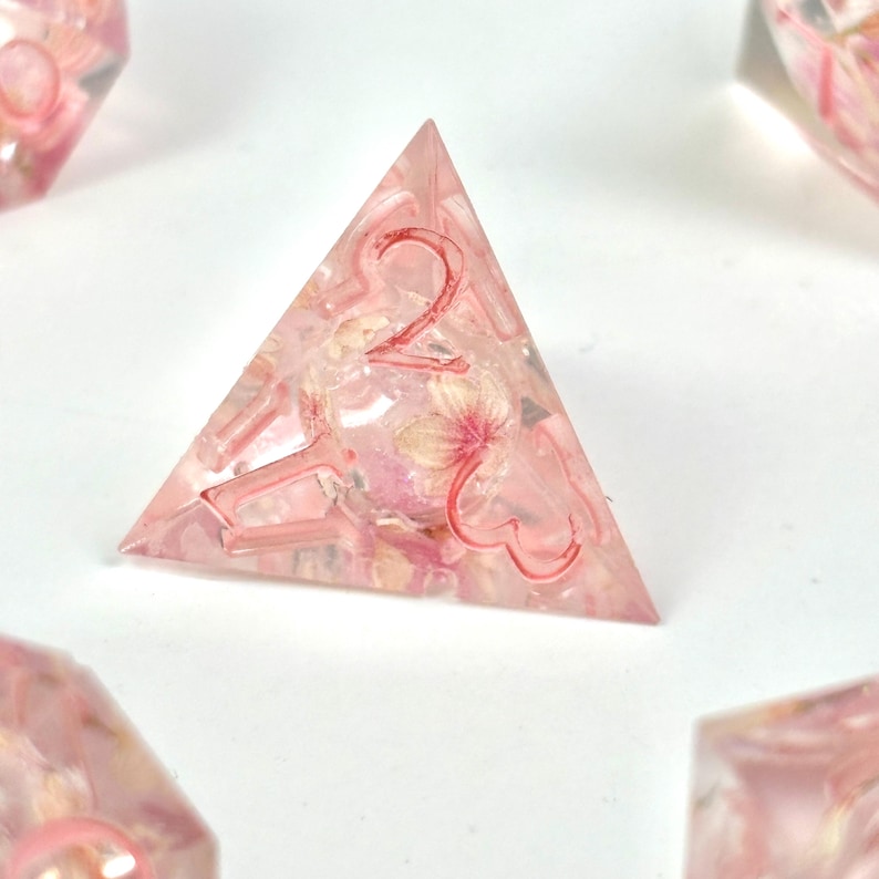 May include: A pink, triangular resin die with the number 2 in red. The die is filled with small, dried flowers in shades of pink and white. Other dice are visible in the background.