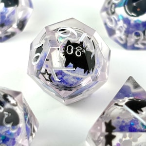 Puede incluir: Un conjunto de dados poliédricos con un diseño transparente y facetado. Cada dado presenta una silueta de gato negro y números blancos. El interior está lleno de purpurina azul y resina de color lavanda.