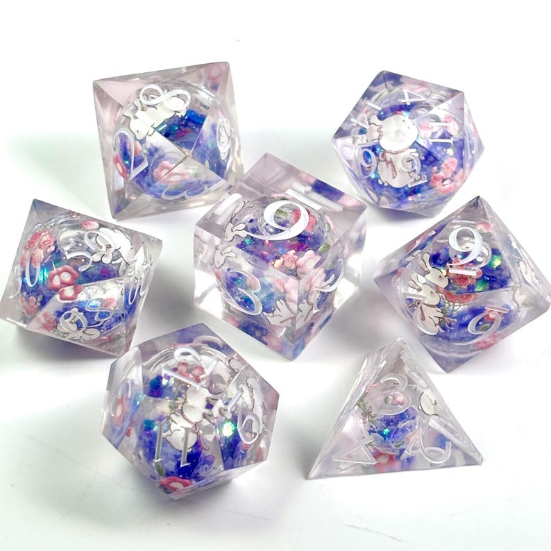 Peut inclure: Ensemble de sept d&eacute;s en r&eacute;sine transparente avec des motifs floraux et de lapins. Les d&eacute;s pr&eacute;sentent des accents bleus, roses et blancs, avec des chiffres blancs. Les d&eacute;s sont de formes g&eacute;om&eacute;triques vari&eacute;es, dont un cube, un d20 et un d4.