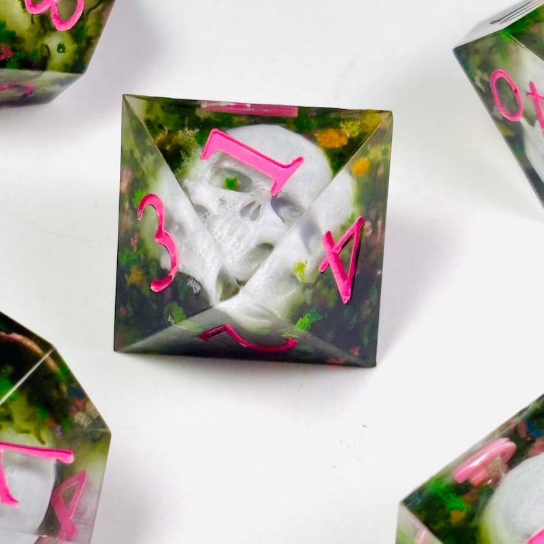 Op de afbeelding: Vierkante dobbelstenen met een schedelontwerp ingekapseld in heldere hars. De dobbelstenen hebben een groene en bloemrijke achtergrond met felroze cijfers. Deze dobbelstenen zijn ontworpen voor tabletop gaming.