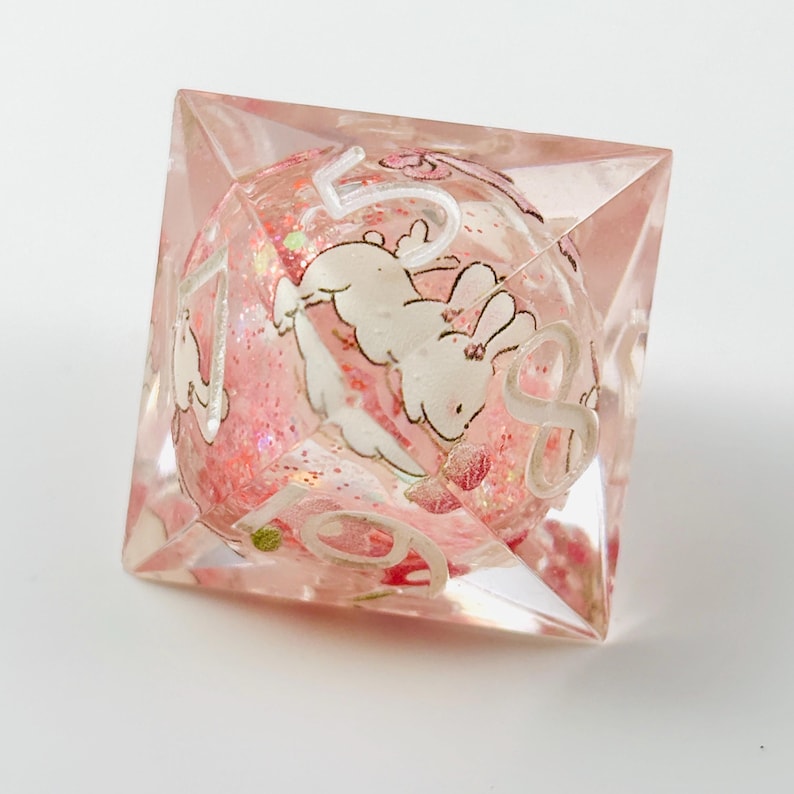 Peut inclure: Un d&eacute; transparent rose &agrave; plusieurs faces avec un c&oelig;ur sph&eacute;rique. Le c&oelig;ur est rempli de paillettes roses et pr&eacute;sente des illustrations de lapins blancs et des chiffres. Le d&eacute; est con&ccedil;u pour les jeux de soci&eacute;t&eacute; et a une esth&eacute;tique fantaisiste.