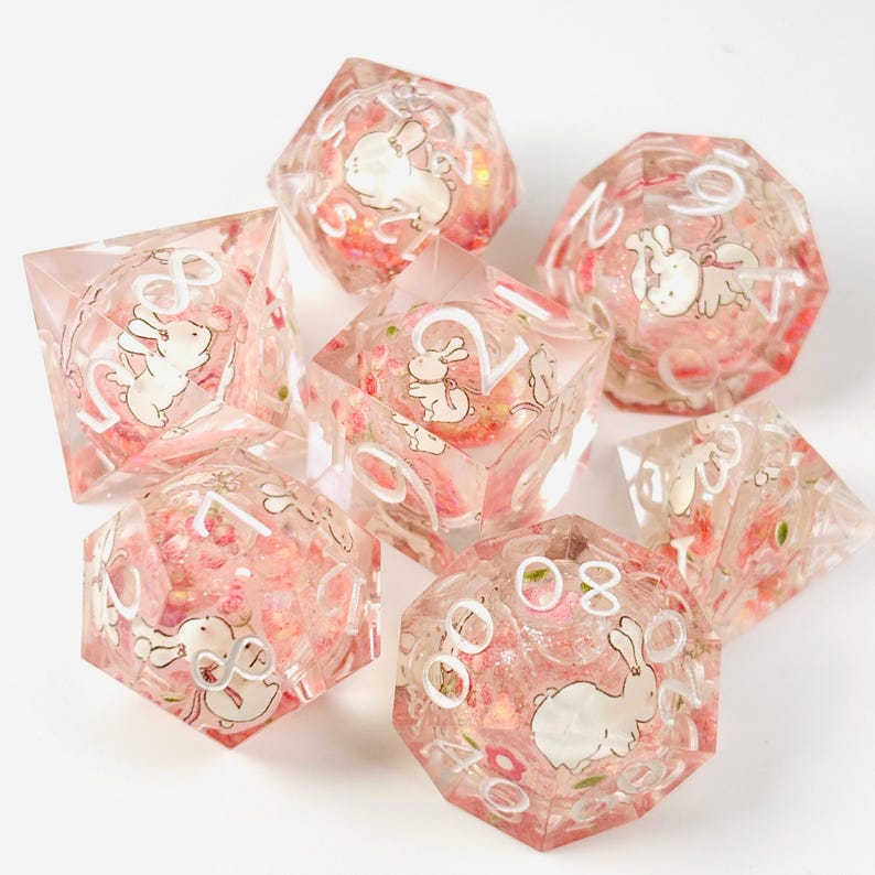 Peut inclure: Ensemble de sept d&eacute;s en r&eacute;sine transparente avec un motif floral rose et des illustrations de lapins blancs. Chaque d&eacute; comporte des chiffres blancs. Ces d&eacute;s sont con&ccedil;us pour les jeux de soci&eacute;t&eacute; et les jeux de r&ocirc;le.